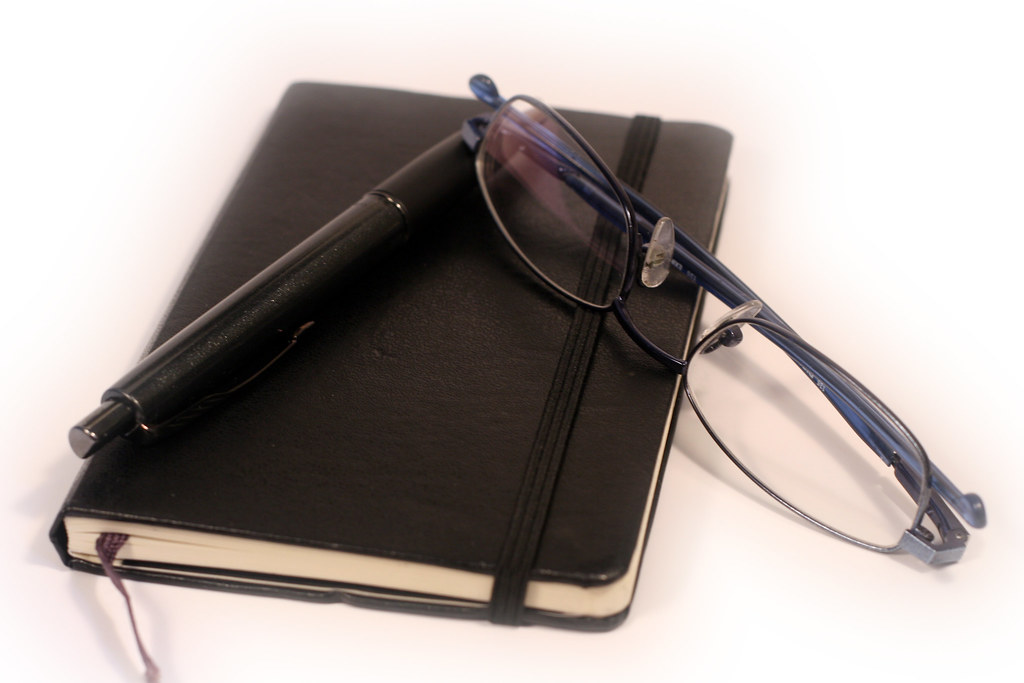 Cuaderno, boligrafo y gafas sobre una mesa, representando la toma de apuntes